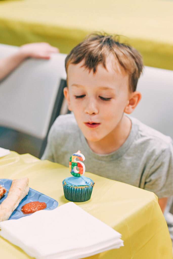 Child blowing out a LEGO-themed birthday candle on a cupcake at a party table